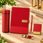 Premium red corporate gift hamper featuring a textured hardcover notebook with golden nameplate closure, matching insulated stainless steel bottle, metallic red pen, personalized visiting card holder and stylish red strap metal keychain, presented in an elegant wooden gift box with cream cushioning.