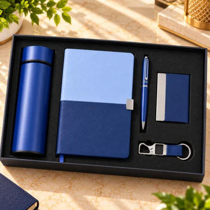 Premium blue personalized corporate gift hamper featuring a dual-tone hardcover notebook, insulated stainless steel bottle, matching metal pen, visiting card holder and customized metal keychain, arranged in a premium black presentation box.