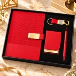 Premium red corporate gift set featuring a textured hardcover personalized notebook with golden nameplate, matching visiting card holder, metallic red pen and customized red strap metal keychain, beautifully arranged in a luxury black presentation box.